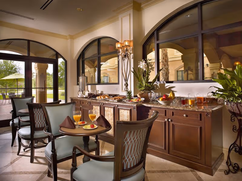 A cozy dining area with a small round table set for two, featuring two glasses of orange juice, plates with fruit, and folded brown napkins. Behind the table is a long wooden buffet with a marble countertop displaying various breakfast items, including pastries, fruit, and pitchers of juice. Large arched windows and doors provide a view of an outdoor patio with tables and umbrellas. The room is warmly lit with wall sconces and decorated with plants and flowers.