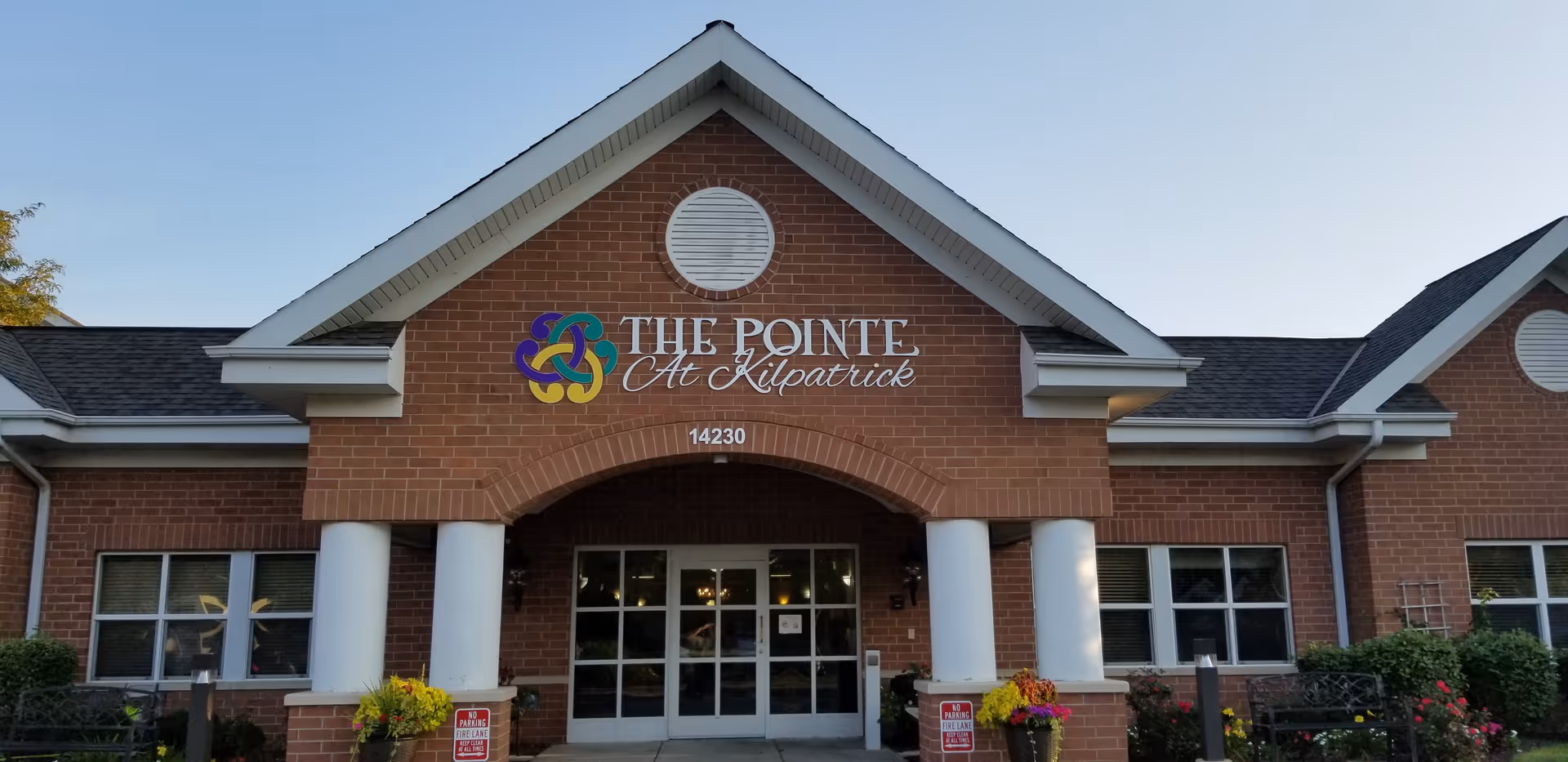 Front exterior view of a brick building with white columns and a peaked roof. The building has a sign that reads 'THE POINTE At Kilpatrick' above the entrance doors. There are plants and flowers near the entrance and two signs indicating no parking in the fire lane.