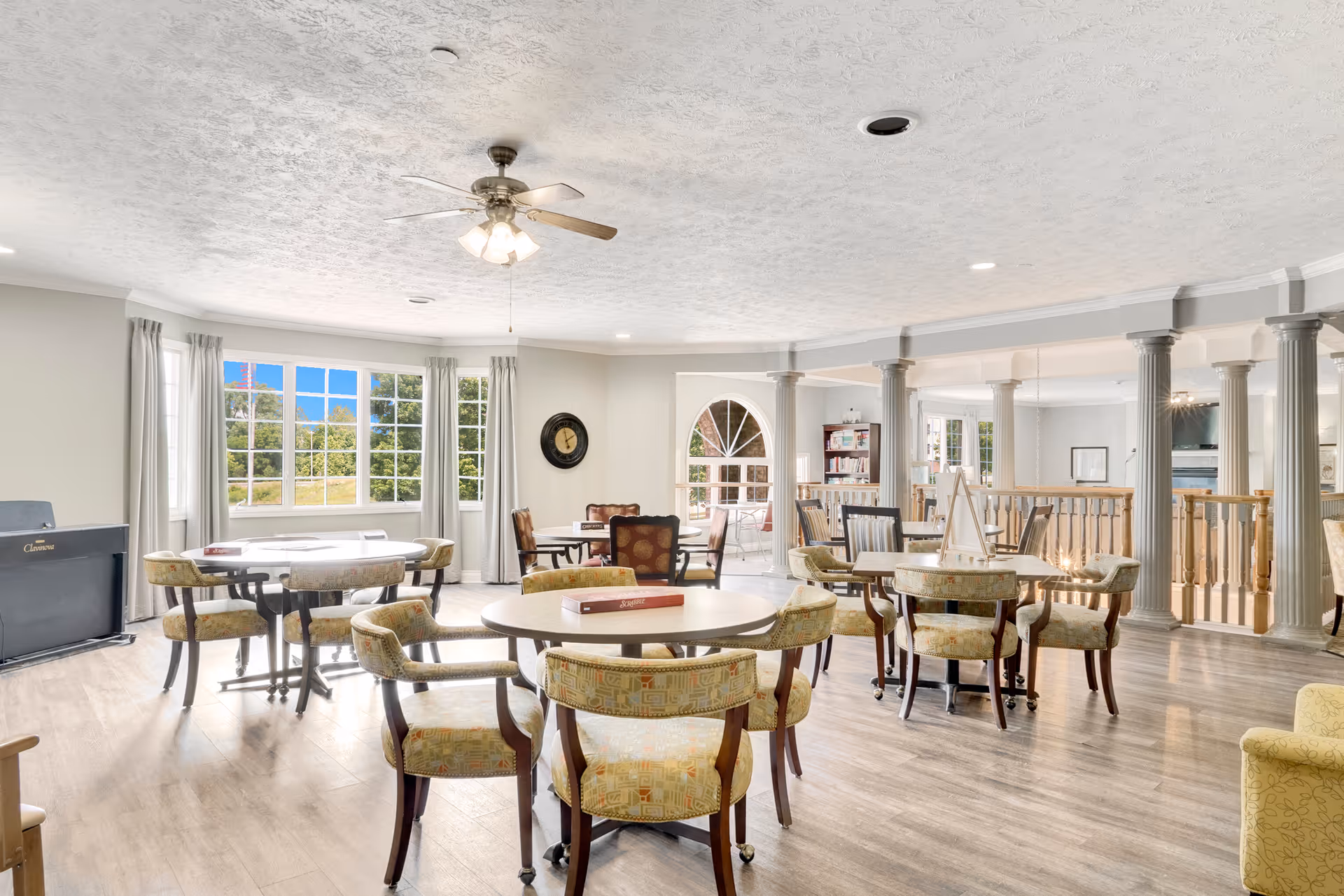 A bright and spacious common area with multiple round tables and cushioned chairs arranged for group seating. Large windows with light curtains allow natural light to fill the room, and a ceiling fan with lights hangs from the textured ceiling. The room features wooden flooring, decorative columns, and a railing separating different sections of the space. A clock is mounted on one wall, and bookshelves and additional seating are visible in the background.