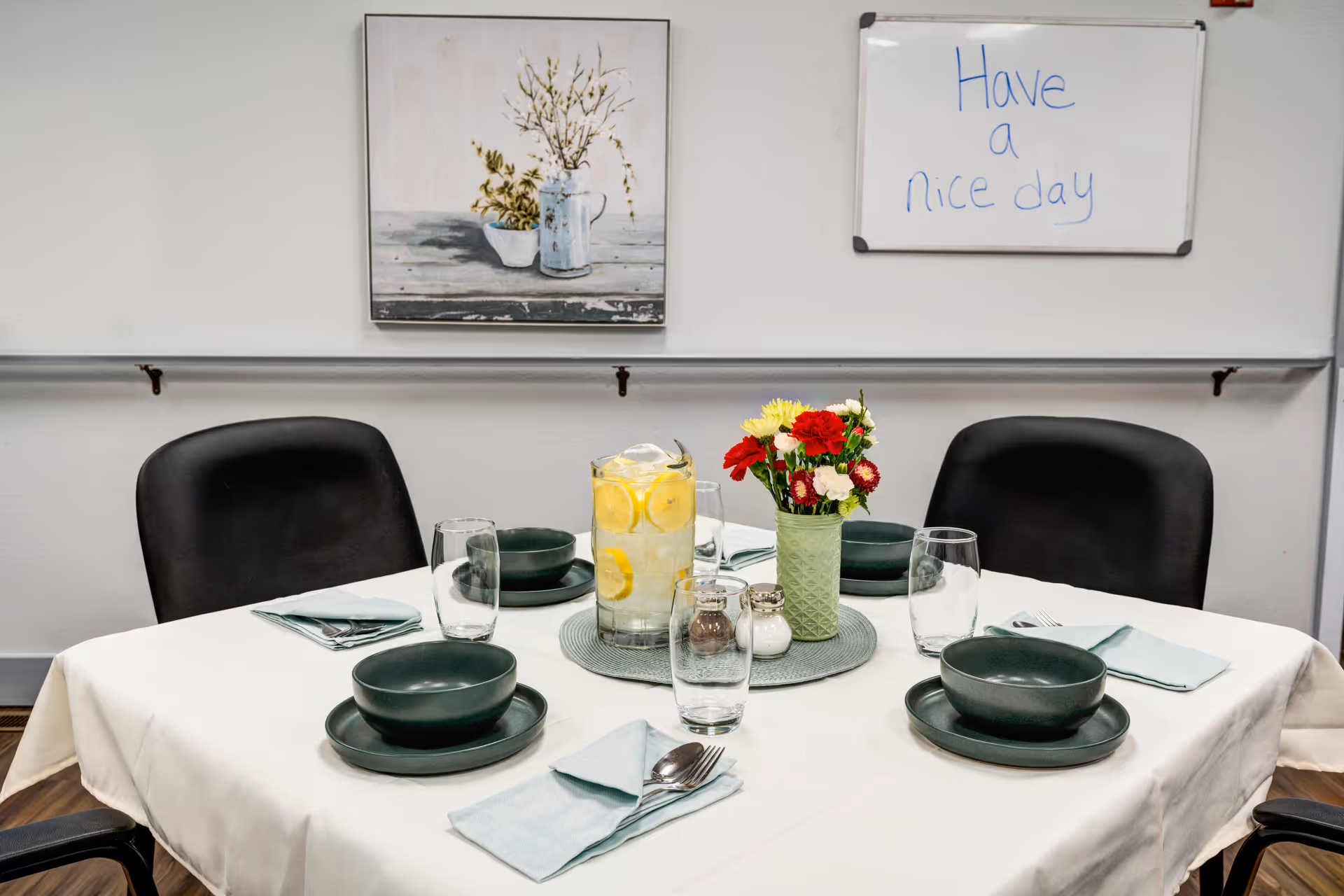 A dining table set for four with dark green bowls and plates, light blue napkins, clear drinking glasses, and silverware. A pitcher of water with lemon slices and a small vase with red and white flowers are placed in the center on a round placemat. Behind the table, there is a painting of a vase with flowers and a whiteboard with the message 'Have a nice day' written in blue marker. The chairs around the table are black and the room has light-colored walls.
