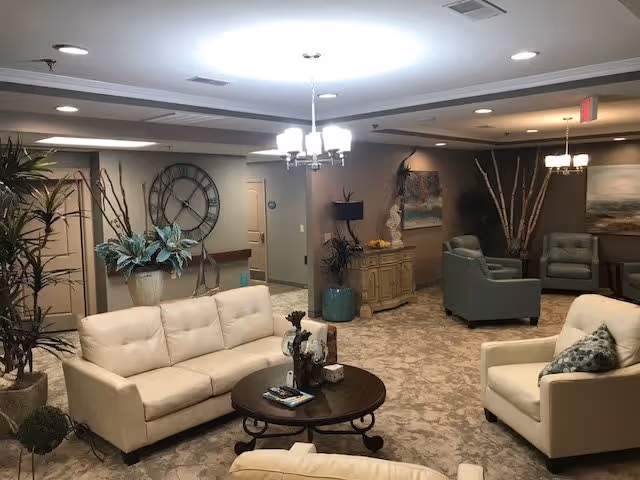 A cozy living room area in a retirement center featuring beige and green upholstered sofas and armchairs arranged around a round wooden coffee table. The room is decorated with plants, a large wall clock, paintings, and soft lighting from ceiling fixtures and lamps. The carpeted floor and neutral wall colors create a warm and inviting atmosphere.