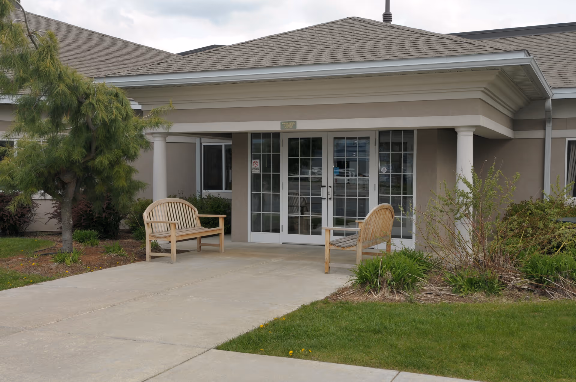 Entrance of a building with double glass doors, two wooden benches on either side of the walkway, surrounded by green grass, shrubs, and a tree under a cloudy sky.