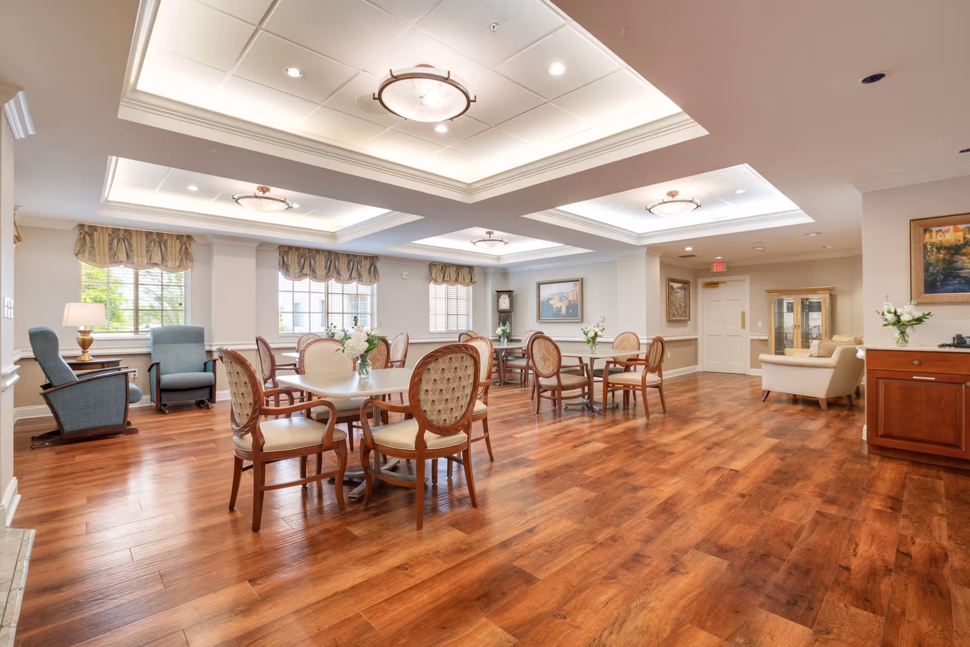 A spacious and well-lit common area in a senior living facility with wooden flooring, several round tables surrounded by cushioned chairs, and a few armchairs near windows with valance curtains. The ceiling features recessed lighting and decorative fixtures, and the walls are adorned with framed artwork and floral arrangements.