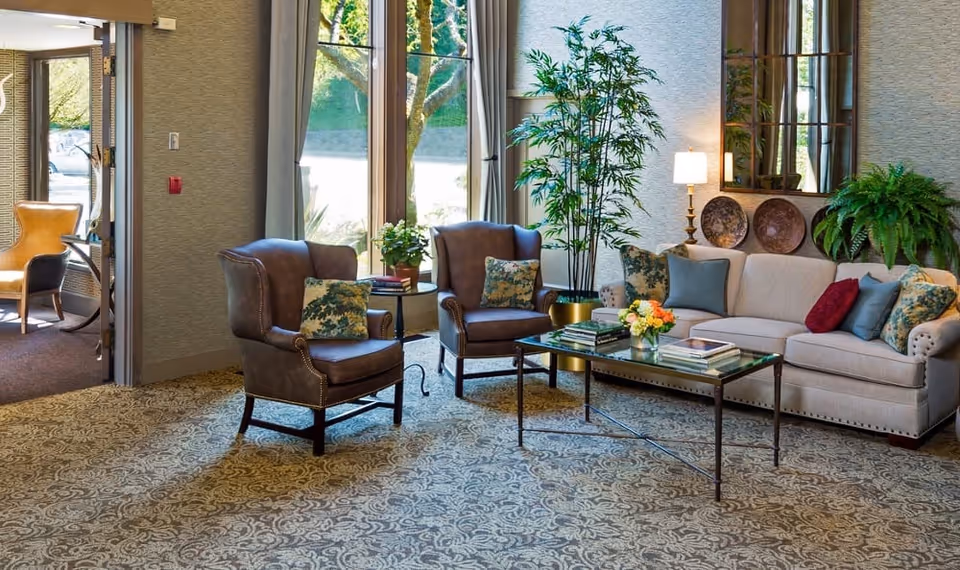 A cozy living room area in Madison House Assisted Living Community featuring a beige sofa with multiple decorative pillows, two brown leather armchairs with patterned cushions, a glass coffee table with books and a flower arrangement, a tall green plant in a gold pot, and large windows with curtains letting in natural light.