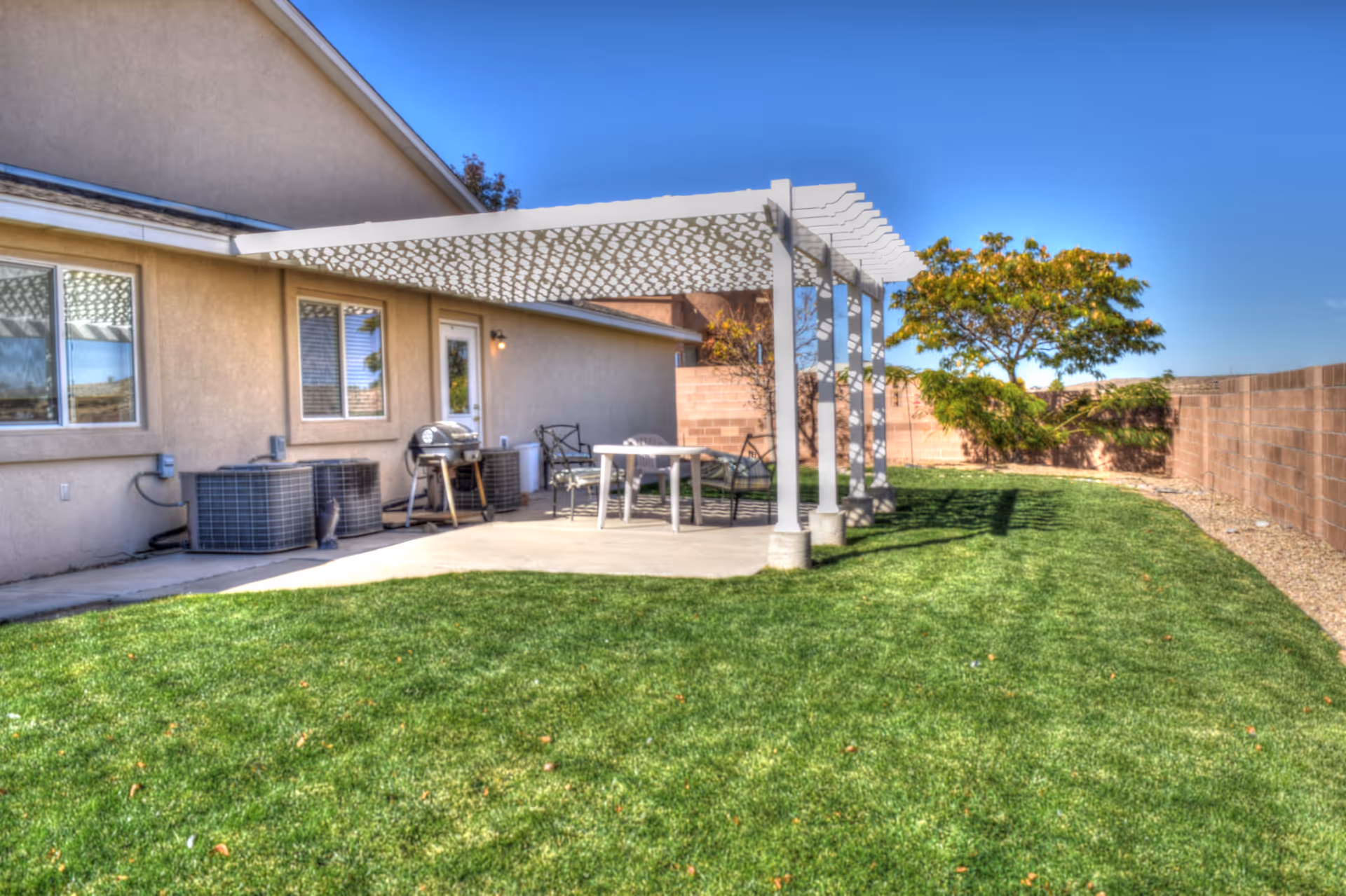Outdoor patio area with a white pergola attached to a beige building. There is a concrete patio with a table, chairs, and a barbecue grill. The patio is surrounded by a well-maintained green lawn and a brick wall fence. A tree and some shrubs are visible near the fence under a clear blue sky.