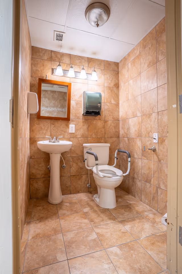 A small bathroom with tan tiled walls and floor, featuring a white pedestal sink with a mirror above it, a toilet with safety rails on both sides, a wall-mounted paper towel dispenser, and a ceiling light fixture.
