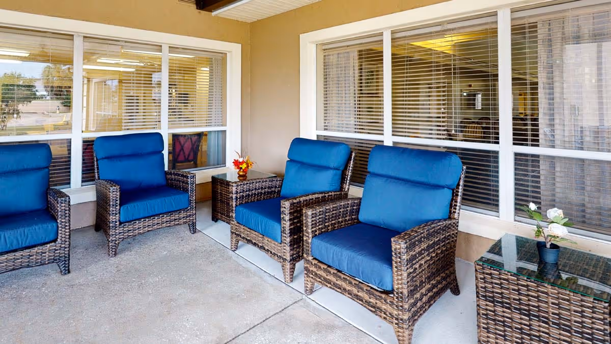 Outdoor seating area with four wicker chairs featuring blue cushions arranged around two wicker side tables, one with a small flower arrangement and the other with a potted plant, set against a beige wall with large windows.