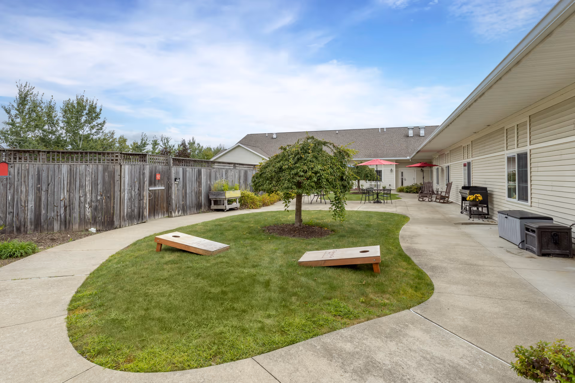 Outdoor courtyard area with a small tree in the center of a grassy patch, two cornhole boards on the grass, concrete walkways surrounding the grass, patio tables with red umbrellas, rocking chairs, a grill, and storage containers along the building wall under a partly cloudy sky.