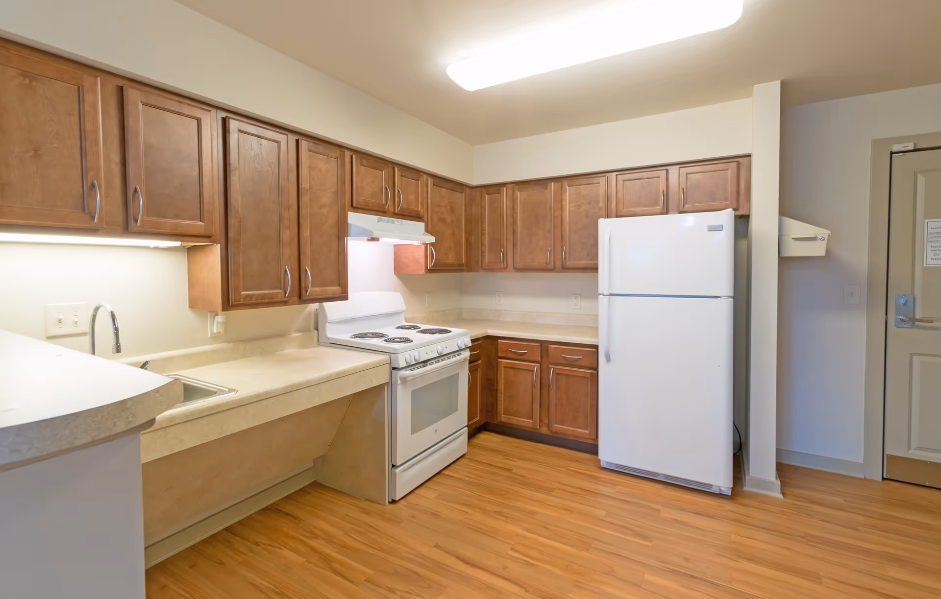 A clean kitchen with wooden cabinets, a white electric stove with oven, a white refrigerator, a sink with a faucet, and beige countertops. The floor is wooden, and there is a door visible on the right side of the image.