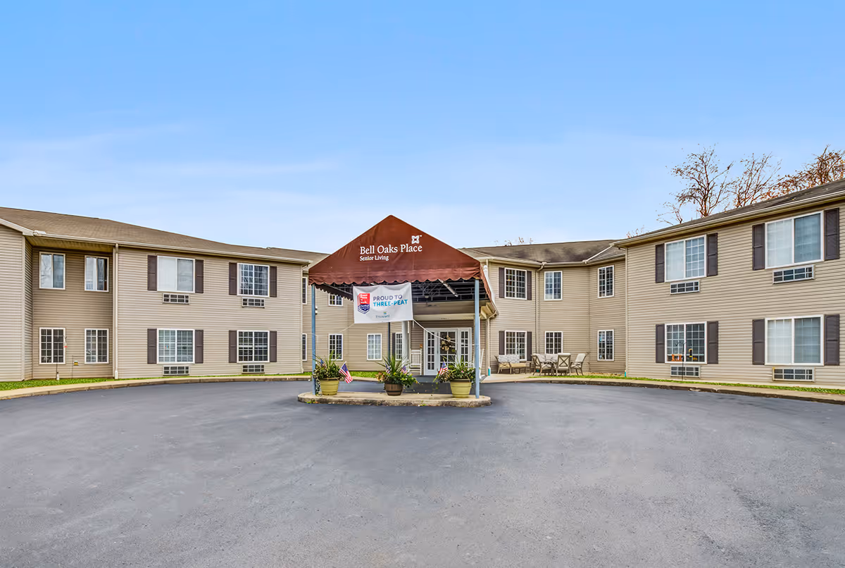 Front exterior view of Bell Oaks Place senior living facility with a circular driveway and a maroon canopy entrance. The building is two stories with beige siding and multiple windows. There are potted plants and American flags near the entrance.