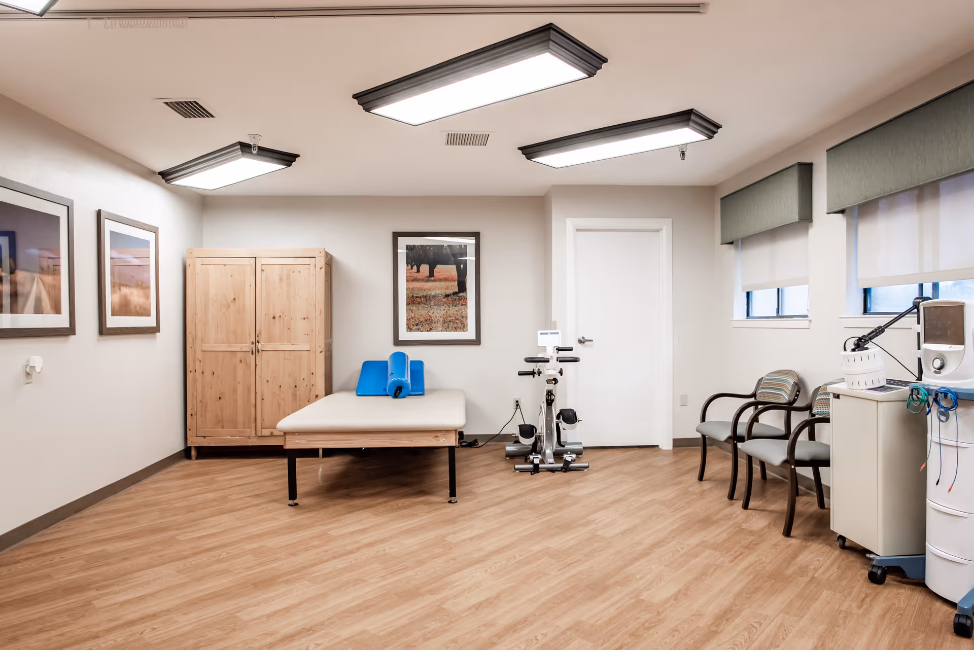 A rehabilitation therapy room with a padded treatment table, blue foam supports, an exercise bike, two chairs, a wooden cabinet, medical equipment on a cart, and framed pictures on the walls. The room has wood flooring, white walls, and windows with green valances and white blinds.