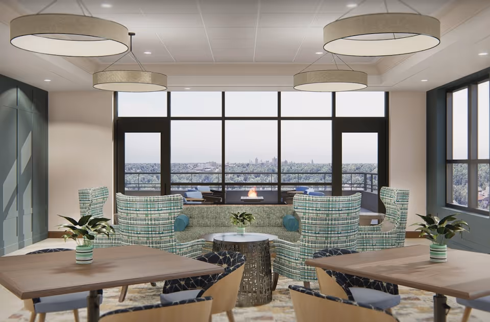 Bright communal lounge with patterned armchairs, tables, pendant lights, and large windows overlooking a city view.