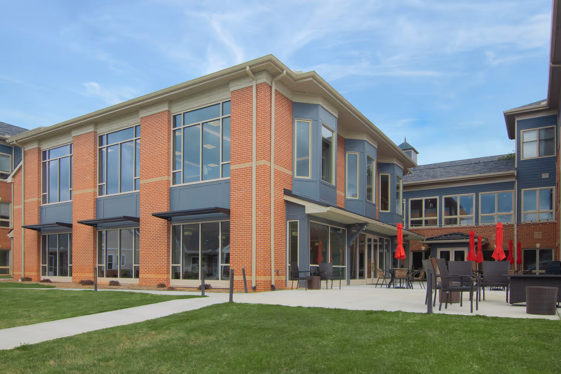 Brick-and-glass two-story retirement community building with a patio, outdoor tables and red umbrellas on a grassy lawn.