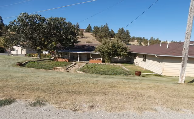 Single-story building with a brown roof and beige siding surrounded by a grassy area and trees, with a hill and more trees in the background under a clear sky.