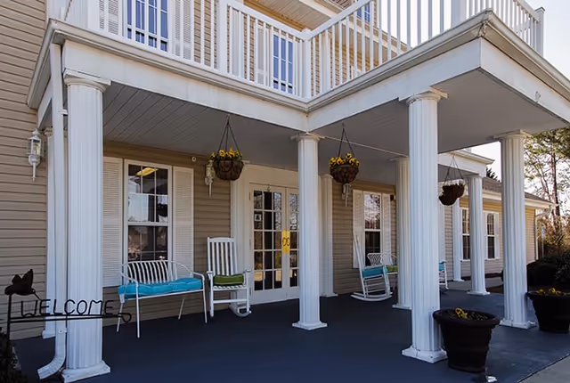 Front porch area of a building with white columns, hanging flower baskets, white rocking chairs with blue cushions, and a welcome sign. The porch has a dark floor and beige siding with white trim around the windows and doors.