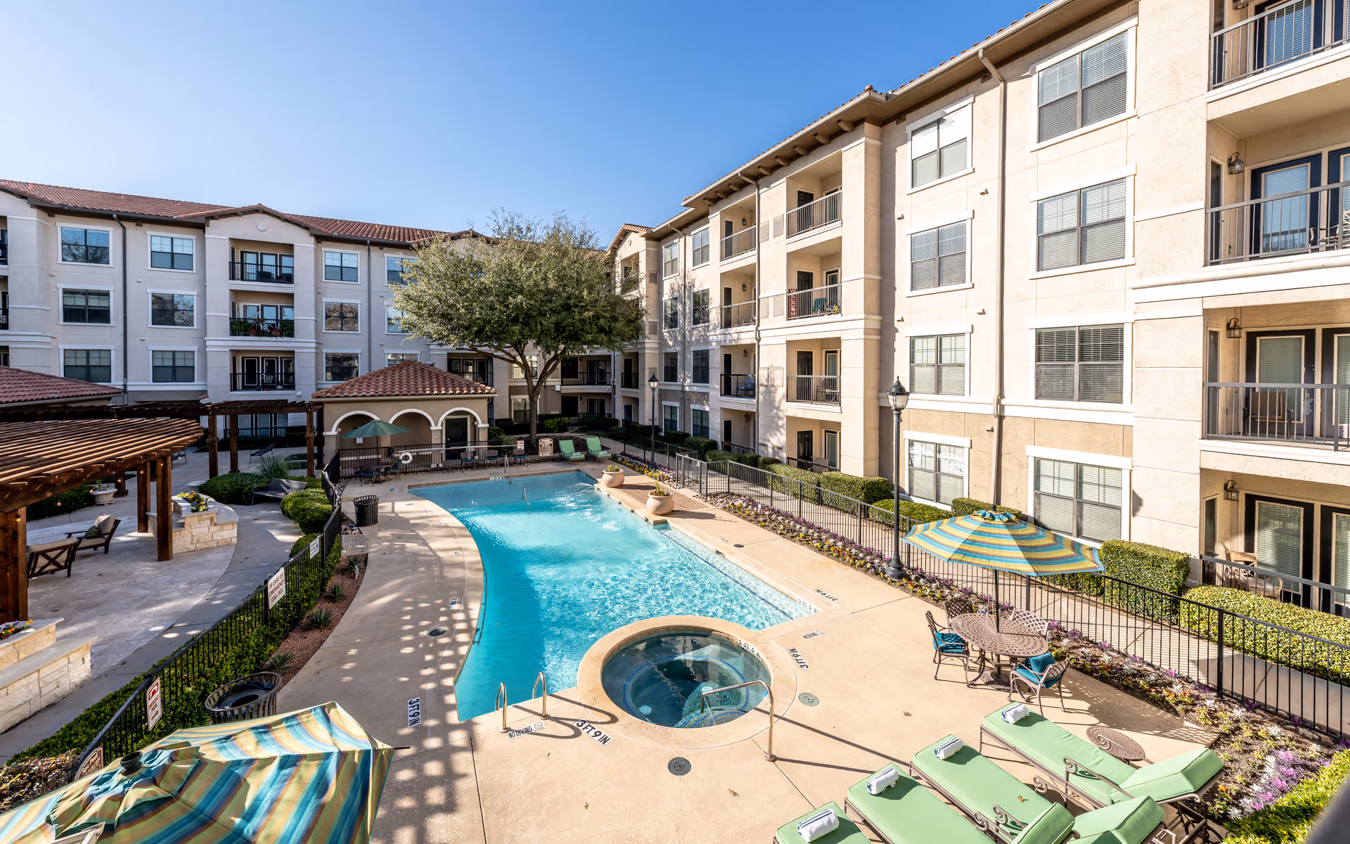 Outdoor swimming pool area surrounded by a multi-story residential building with balconies. The pool area includes a hot tub, lounge chairs with towels, tables with striped umbrellas, and a shaded seating area with pergolas. There are plants and flowers along the poolside and a clear blue sky above.