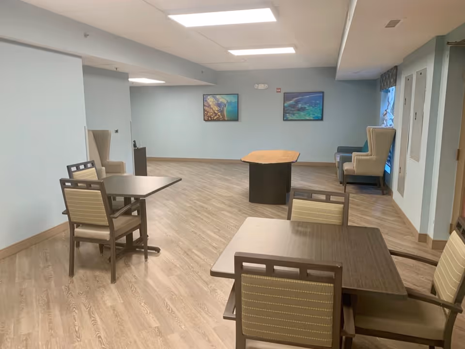 A spacious room with light blue walls and wood-patterned flooring featuring several tables and chairs arranged for seating. Two framed pictures hang on the far wall, and there are two armchairs and a small couch near a window on the right side. The room is well-lit with ceiling lights.