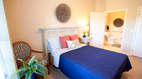 Bright furnished bedroom with a bed covered in a blue quilt, a nightstand and armchair, and an open door revealing an en-suite bathroom.