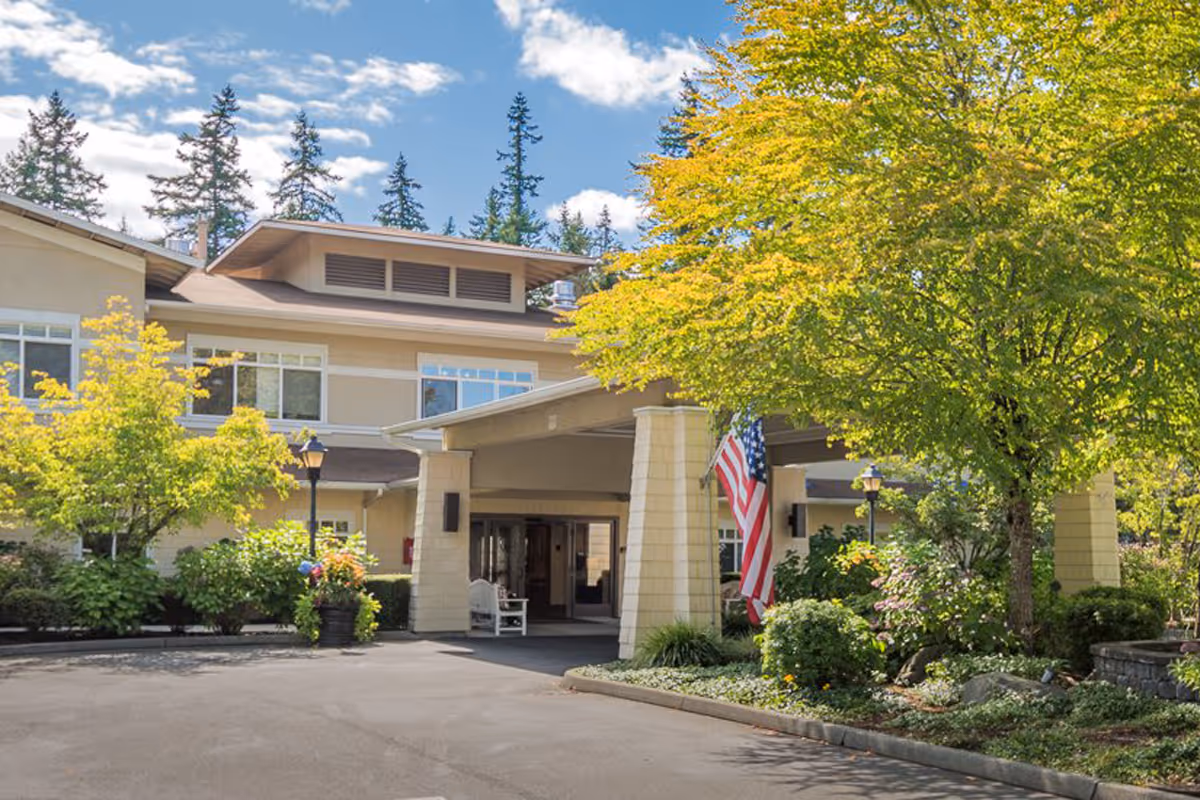 Exterior view of a senior living facility building with a covered entrance, surrounded by trees and landscaping under a partly cloudy sky. An American flag is displayed near the entrance.