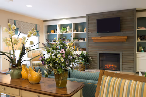 Cozy living room with a fireplace and TV, built-in shelves, floral arrangements on a console table, and upholstered seating.