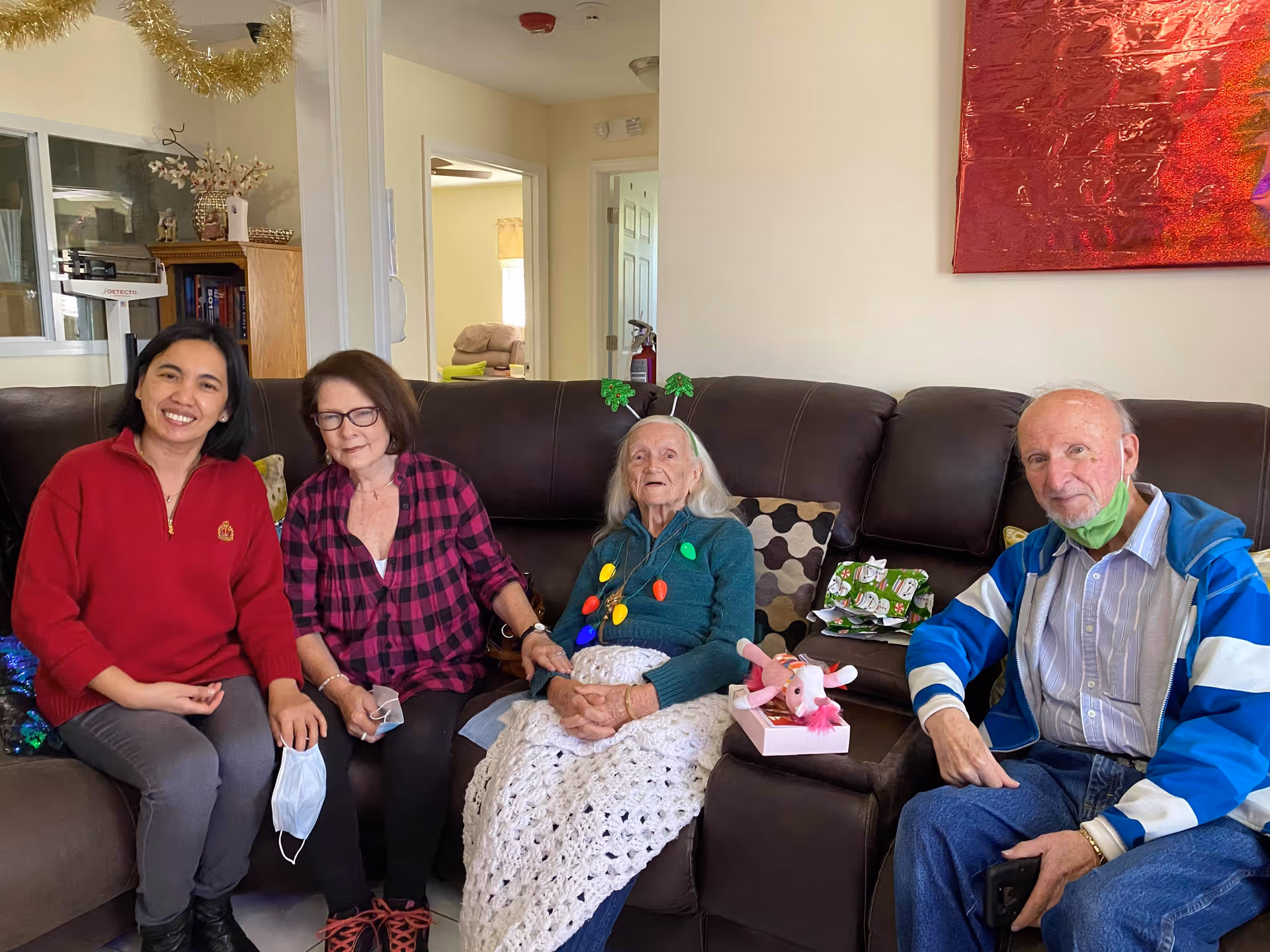 Four people sitting on a brown leather couch in a living room. From left to right: a woman in a red sweater smiling, a woman in a red and black checkered shirt holding a face mask, an elderly woman wearing a green sweater with colorful Christmas lights decoration and a white crocheted blanket on her lap, and an elderly man in a blue and white jacket with a green face mask under his chin. There are holiday decorations visible, including a gold garland hanging from the ceiling and wrapped gifts on the couch. The background shows an open doorway and part of the living space.