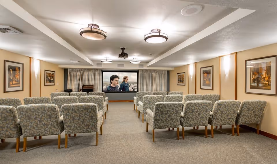 A small theater room with three rows of floral-patterned armchairs facing a large screen displaying a movie scene. The room has beige walls adorned with framed artwork, ceiling lights, and curtains behind the screen.