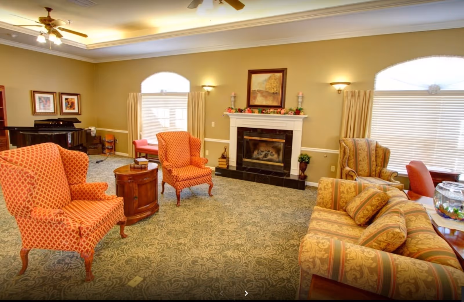 A cozy senior living facility common area featuring patterned armchairs and a sofa arranged around a wooden side table. The room has a fireplace with decorative items on the mantel, two large arched windows with blinds and curtains, a piano in the corner, framed artwork on the walls, and ceiling fans with lights.