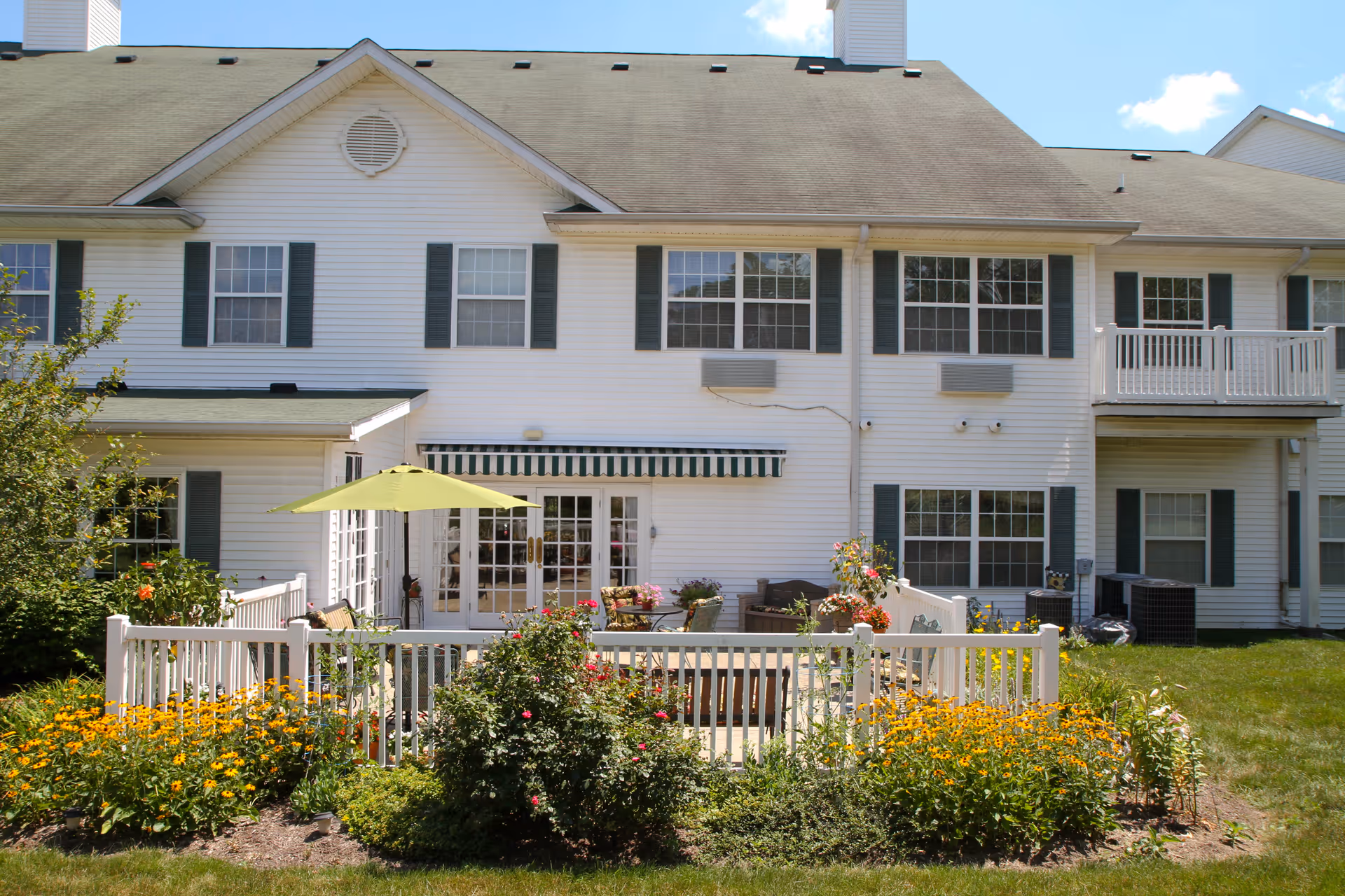 Exterior view of a white two-story residential building with multiple windows and green shutters. The ground floor features a fenced patio area with outdoor furniture, a green umbrella, and colorful flower beds surrounding the patio. The sky is clear with a few clouds.