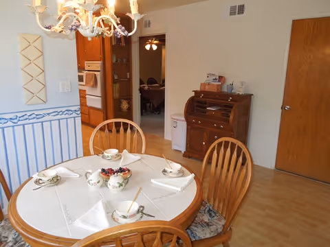 A dining area with a round table set for four, featuring floral-patterned teacups, saucers, and napkins. The table is surrounded by wooden chairs with cushioned seats. A chandelier hangs above the table. In the background, there is a wooden roll-top desk, a closed wooden door, and an open doorway leading to another room with a ceiling fan and additional seating. The walls are light-colored with a blue and white striped wallpaper border.