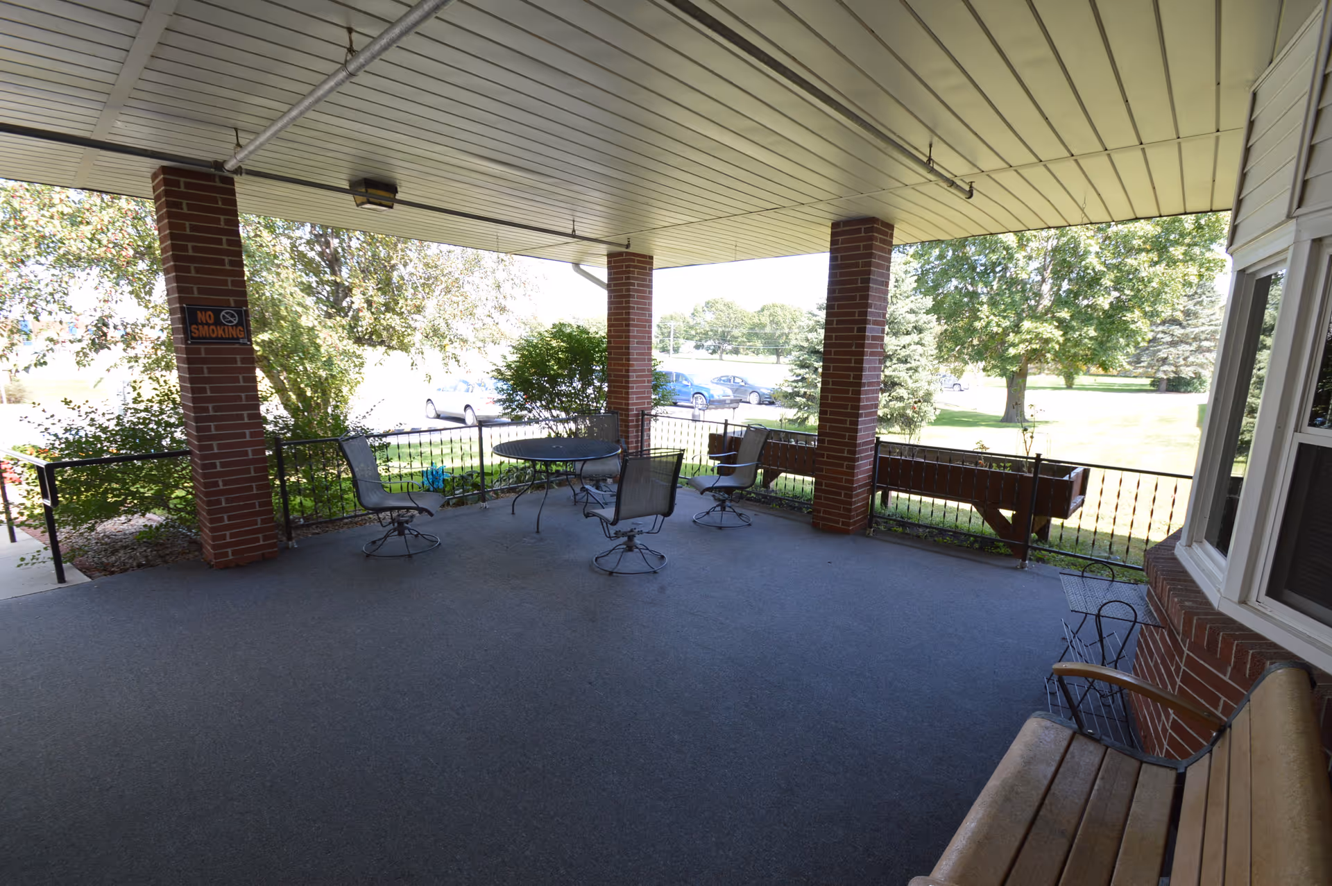 Covered outdoor patio area with a ceiling, brick pillars, and metal railing. There are several metal chairs and a round table arranged on the carpeted floor. A wooden bench is positioned near a window on the right side. Trees and parked cars are visible in the background.