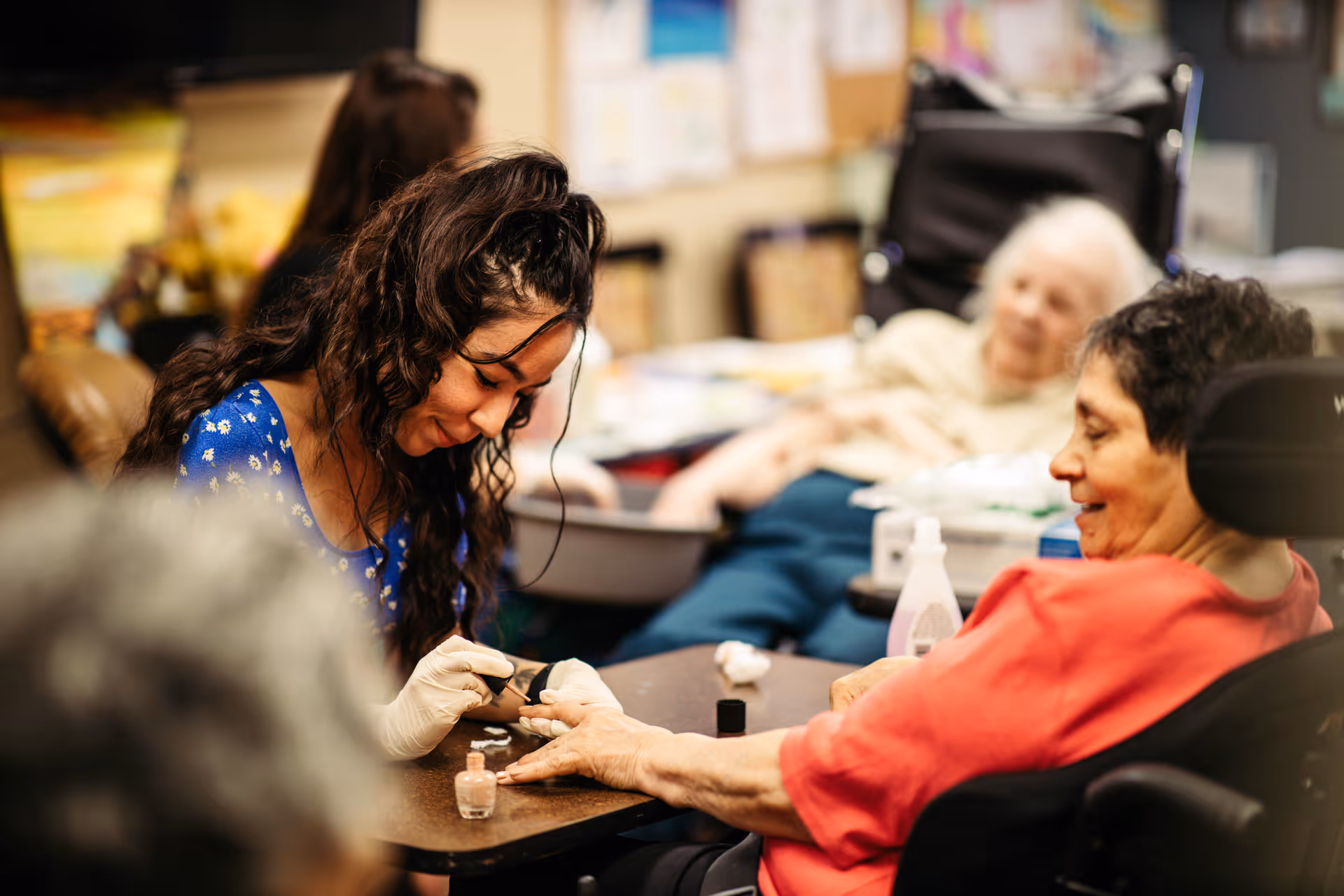 A young woman with gloves is painting the fingernails of an elderly woman seated in a wheelchair, while another elderly woman is visible in the background, all in a cozy indoor setting.