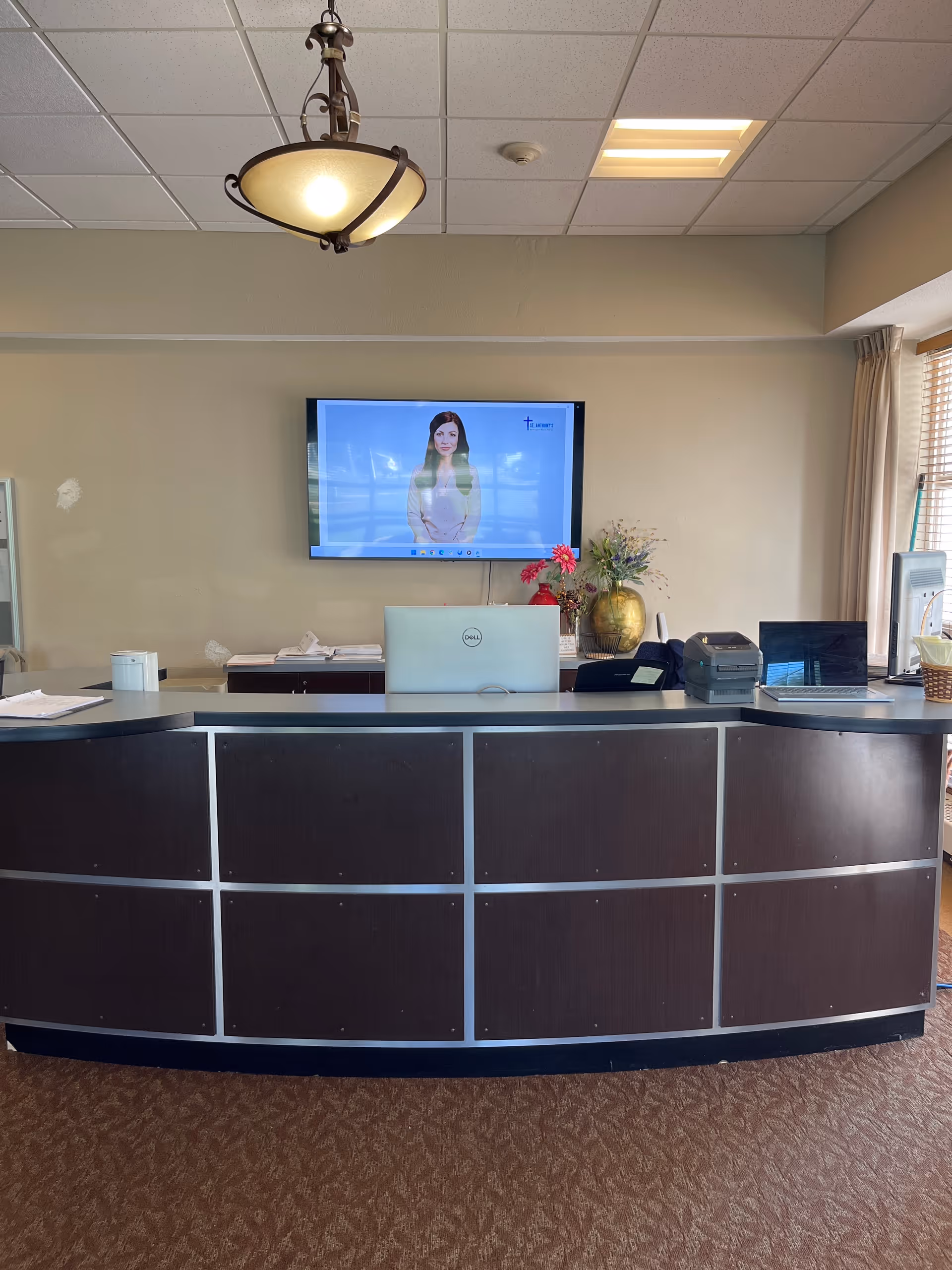 Reception desk area with a curved dark wood front panel and a countertop holding a Dell laptop, a printer, and some papers. Behind the desk is a wall-mounted flat screen TV displaying a woman in a light-colored outfit. A decorative vase with flowers is placed on the counter behind the desk. The room has beige walls, a carpeted floor, a hanging ceiling light fixture, and a window with curtains on the right side.
