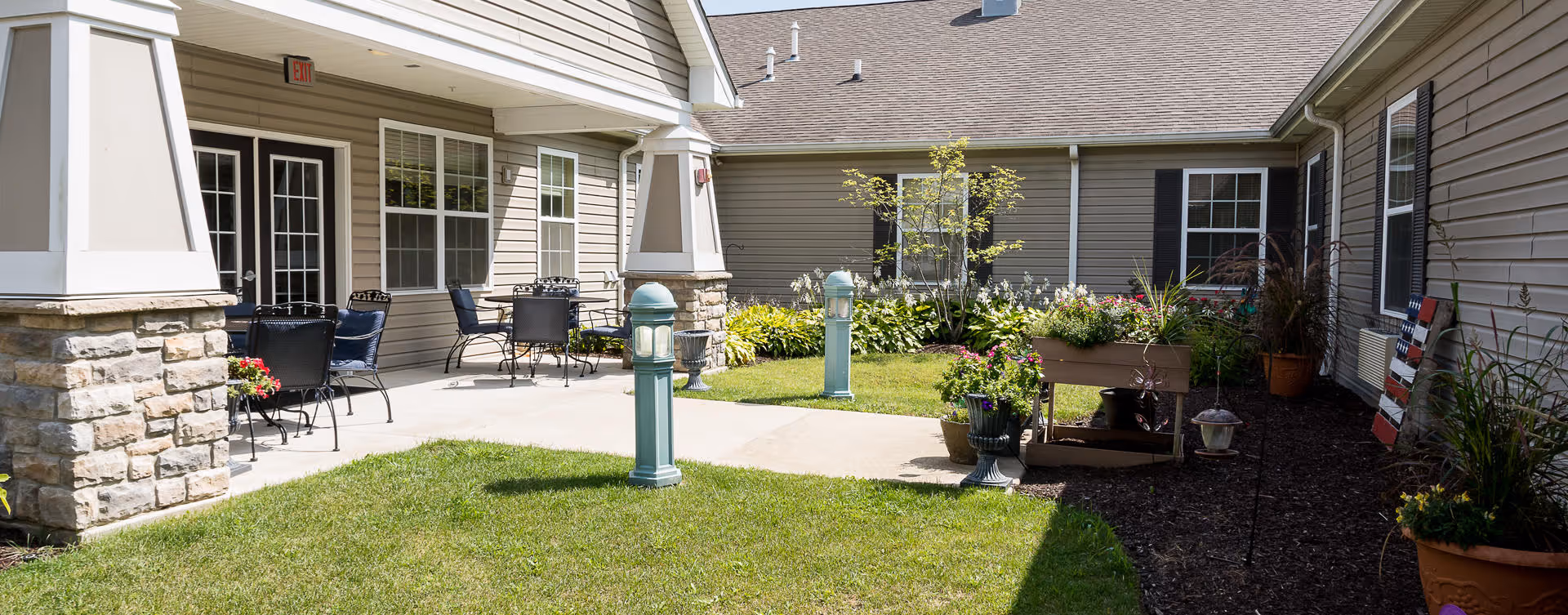 Outdoor courtyard area of a senior living facility with a well-maintained lawn, flower beds, and patio seating including tables and chairs. The building exterior features beige siding and stone pillars supporting a covered walkway.