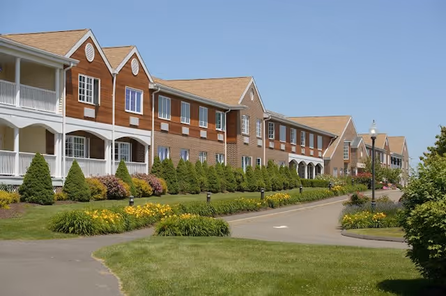 Exterior view of a senior living facility building with multiple peaked roofs, large windows, and white balconies. The building is surrounded by neatly trimmed bushes, green lawns, and flower beds along a paved driveway under a clear blue sky.