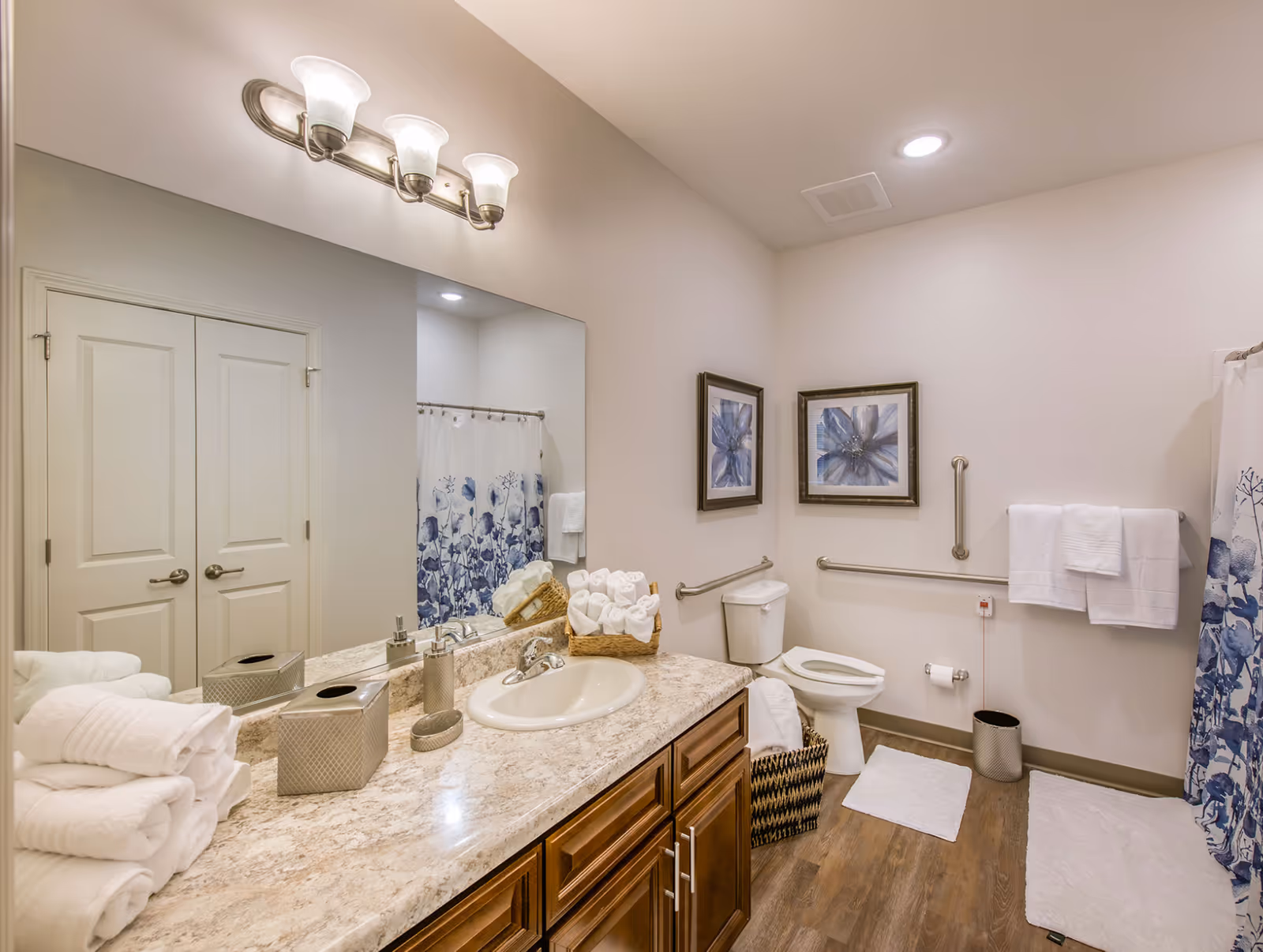 A clean and well-lit bathroom with a large mirror above a marble countertop with a sink. The countertop has neatly folded white towels, a tissue box, and a soap dispenser. The bathroom features a toilet with safety grab bars on the walls, two framed floral pictures above the toilet, a trash can, and a shower with a blue and white floral curtain. The floor is wood-style with white bath mats.