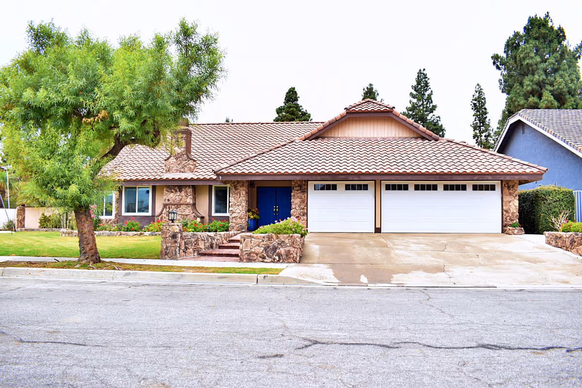 Single-story suburban house with a tile roof, stone facade, blue front door, three-car garage, and a tree-lined front lawn.