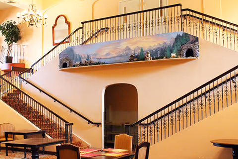 Interior view of a senior living facility featuring a split staircase with decorative wrought iron railings. A mural depicting a scenic mountain landscape with small buildings and trees is painted on the wall above the stair landing. Below the stairs is an arched doorway leading to another room. There are tables and chairs in the foreground, a chandelier hanging from the ceiling, and a large mirror on the wall.