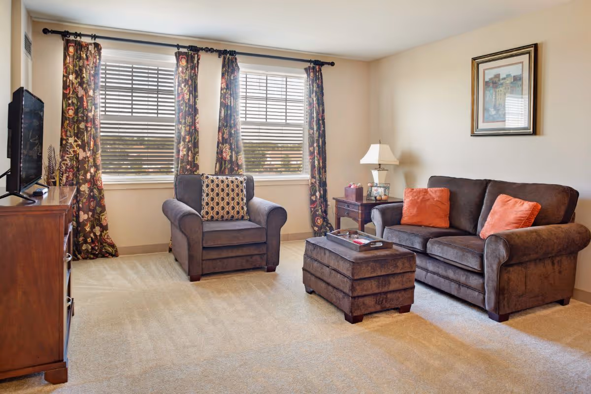 A cozy living room with a sofa, armchair, ottoman, TV console, side table with a lamp, and floral curtains over a large window.