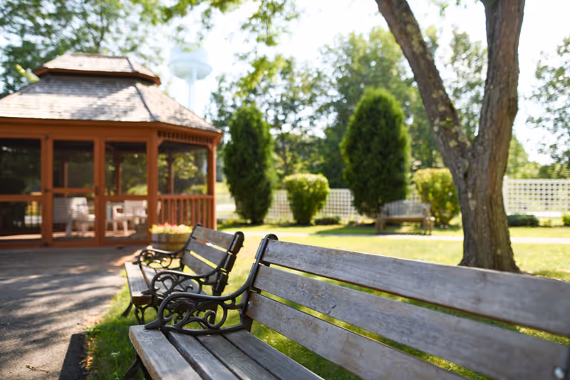 Outdoor area with wooden benches along a paved path, a wooden gazebo, green grass, trees, and shrubs in the background under a sunny sky.