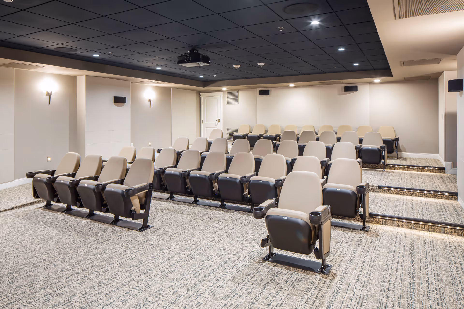 A small theater-style room with several rows of beige and black cushioned seats arranged on a carpeted floor with steps leading up to the back rows. The walls are light-colored with sconces providing soft lighting, and a projector is mounted on the black ceiling.