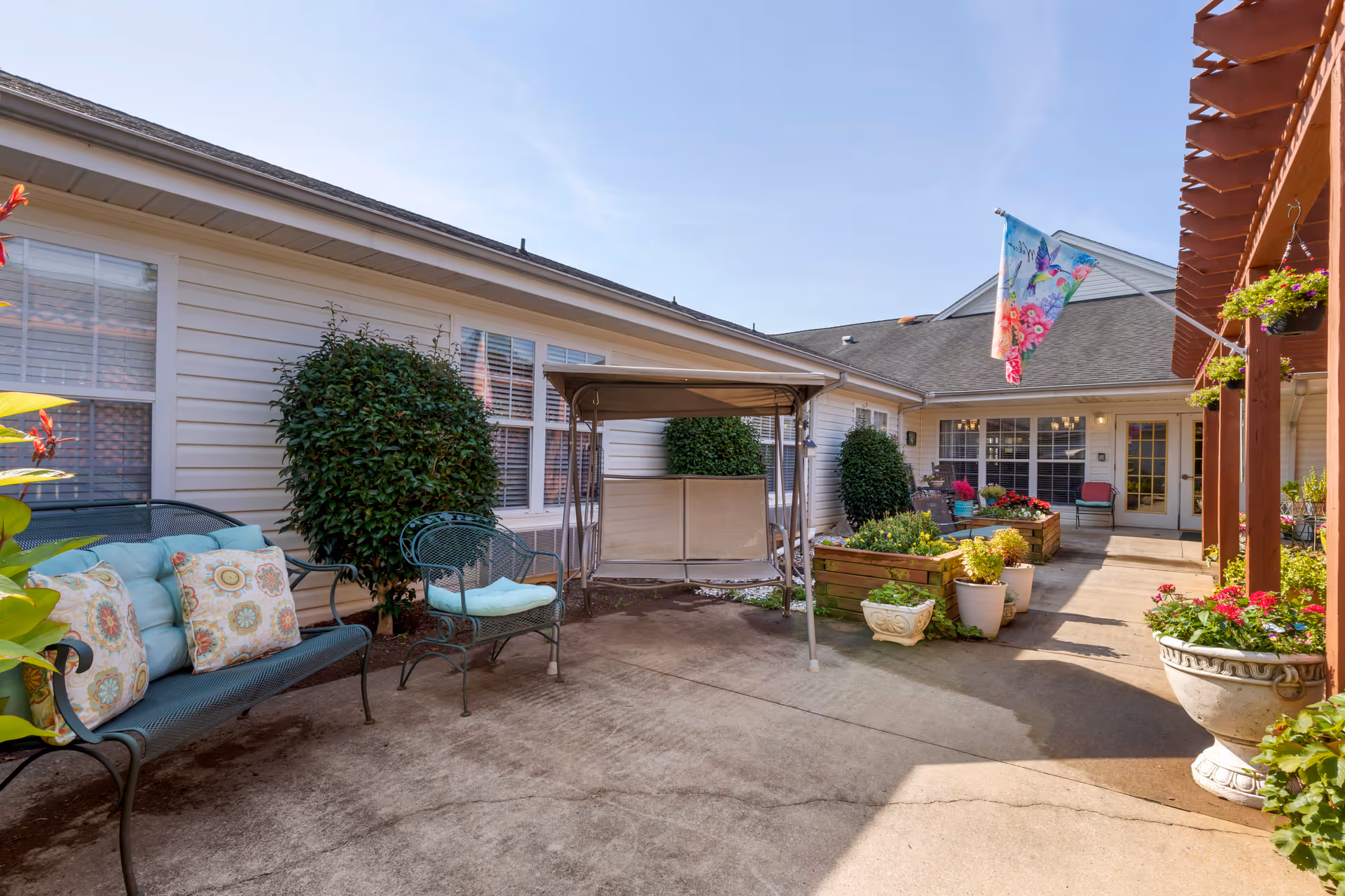 Outdoor patio area at Brookdale Forest City featuring cushioned metal benches, a swing seat, potted plants, flower beds, and a colorful flag hanging from a pergola under a clear blue sky.