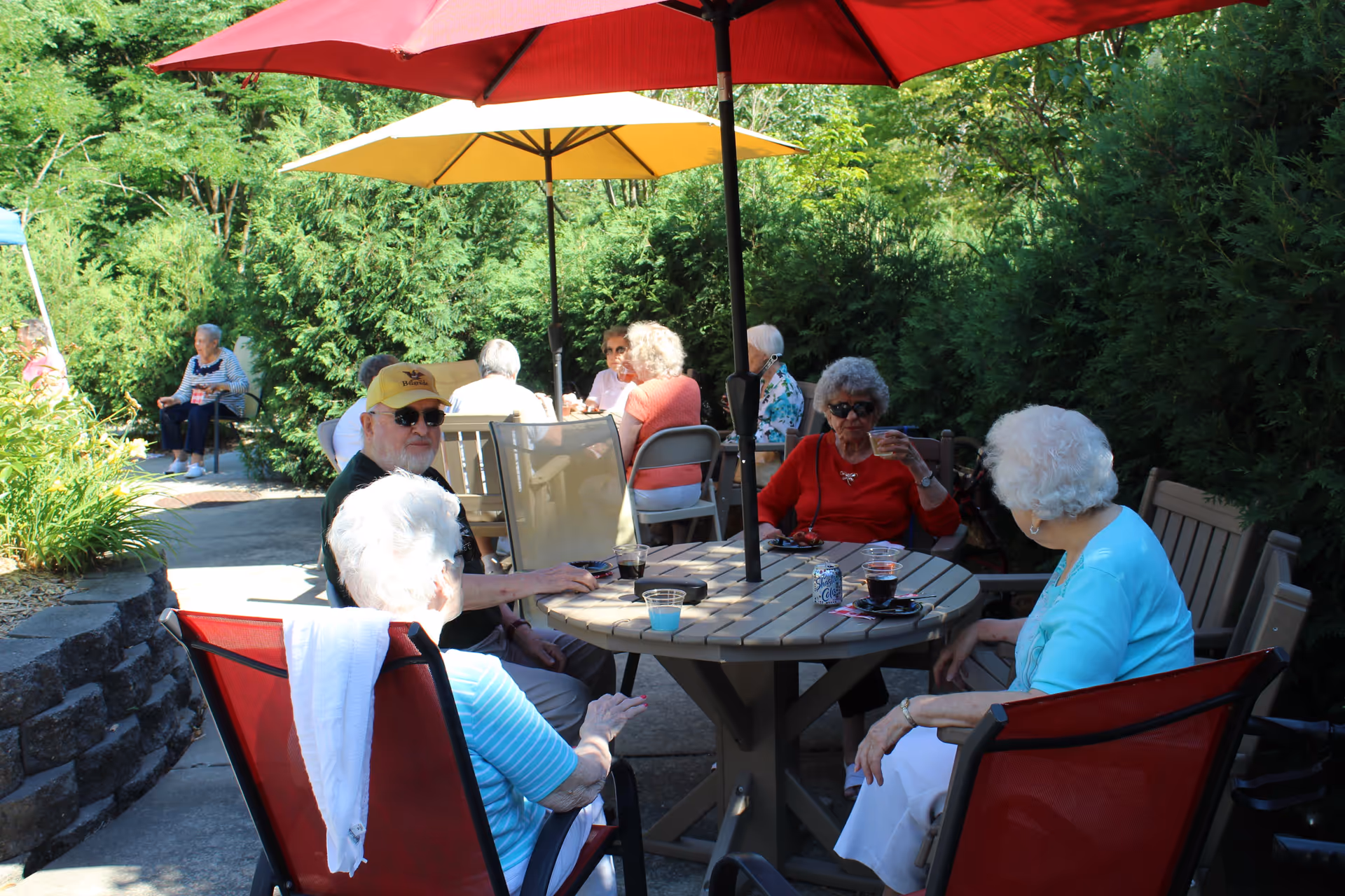 A group of elderly people sitting around a round outdoor table under large red and yellow umbrellas, enjoying drinks and conversation in a garden area surrounded by greenery.