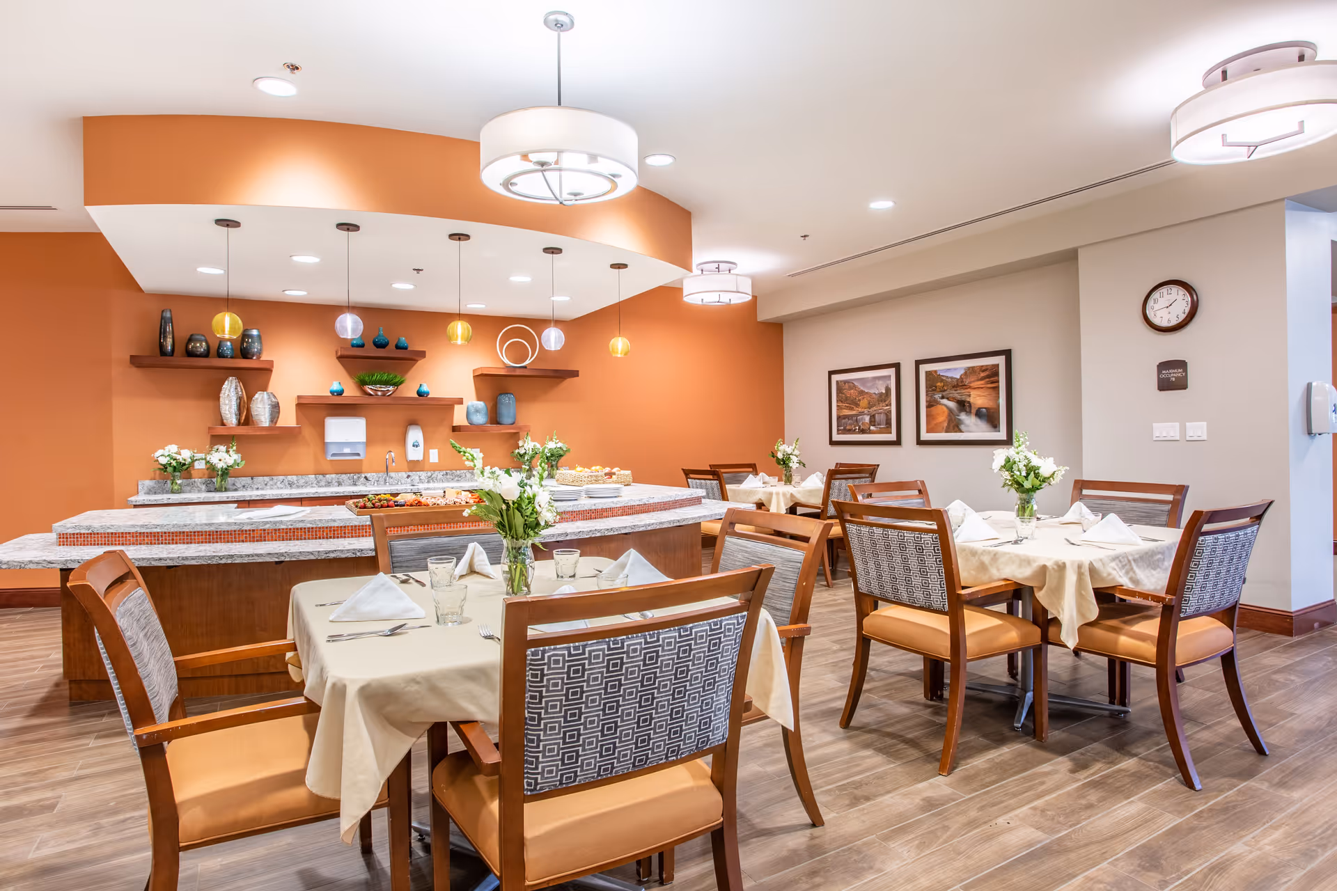 A bright dining room with several tables covered in beige tablecloths, each set with napkins, glasses, and floral centerpieces. The room features wooden chairs with patterned backs and orange cushions. In the background, there is a counter with decorative shelves holding vases and bowls against an orange accent wall. Modern pendant lights hang from the ceiling, and framed landscape pictures are on the adjacent wall.
