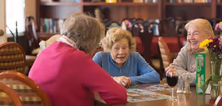 Three elderly women sitting around a table in a dining area, working on a jigsaw puzzle together. One woman is wearing a pink sweater, another a blue sweater, and the third a light-colored sweater. The background shows wooden chairs and tables in a warm, well-lit room.