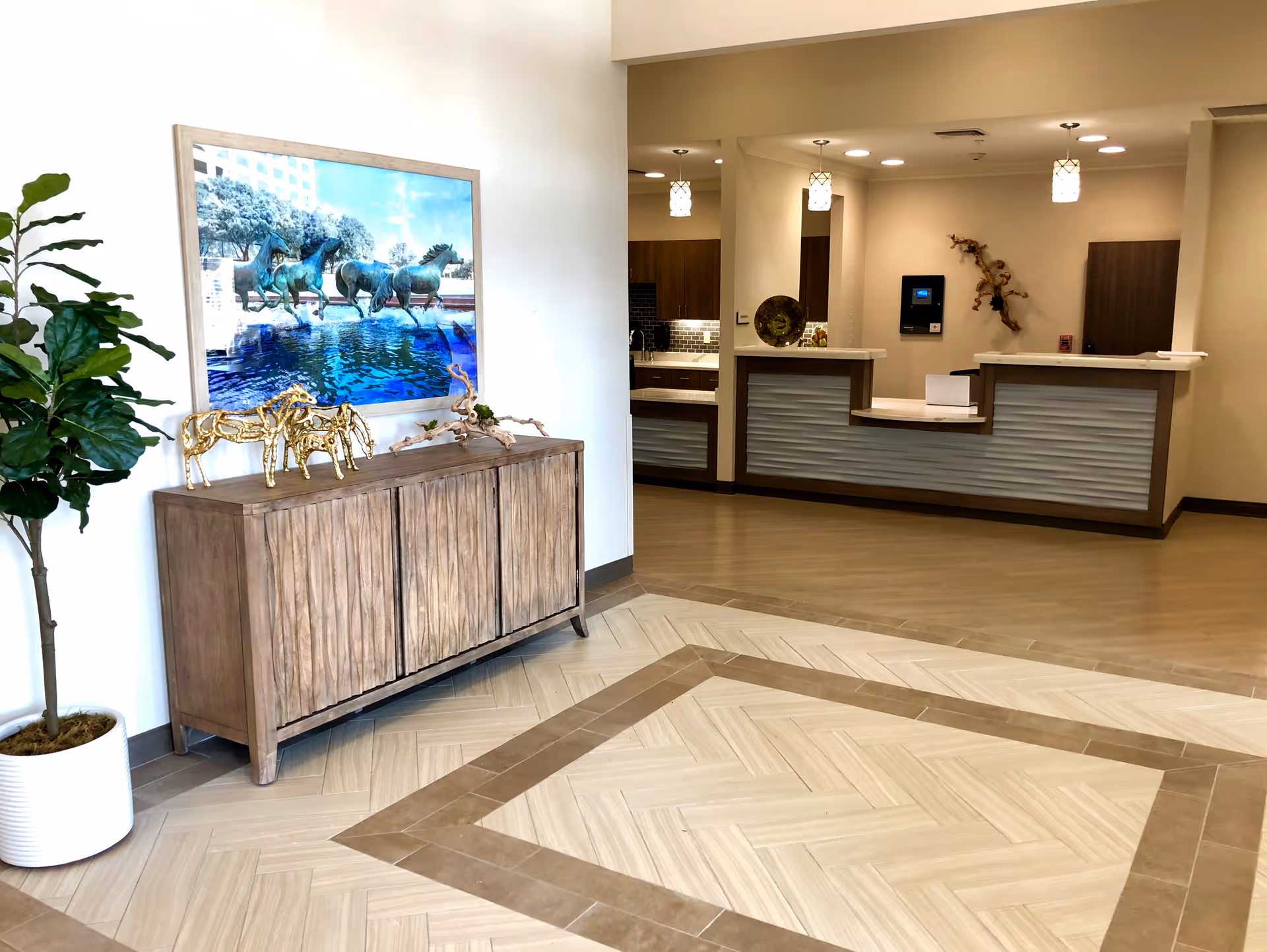Interior view of a reception area in a senior living facility featuring a wooden cabinet with decorative golden horse sculptures and a large framed picture of horses running through water above it. A potted plant is placed to the left of the cabinet. The reception desk is visible in the background with pendant lights hanging from the ceiling and a decorative wall piece behind the desk.
