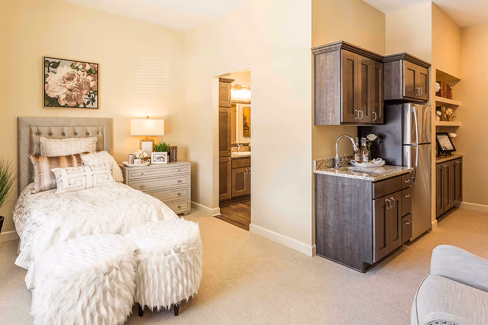 A cozy assisted living room featuring a single bed with multiple pillows and a floral painting above it. Next to the bed is a nightstand with a lamp, books, and decorative items. To the right, there is a small kitchenette with dark wood cabinets, a granite countertop, a sink, and a stainless steel refrigerator. The room has beige walls and carpeted flooring, with a doorway leading to a bathroom visible in the background. Two fluffy white ottomans are placed at the foot of the bed.