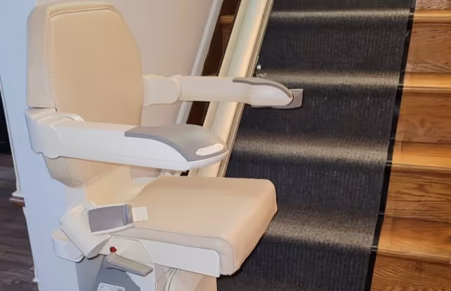Beige stairlift chair mounted on a rail alongside wooden stairs with a dark carpet runner.