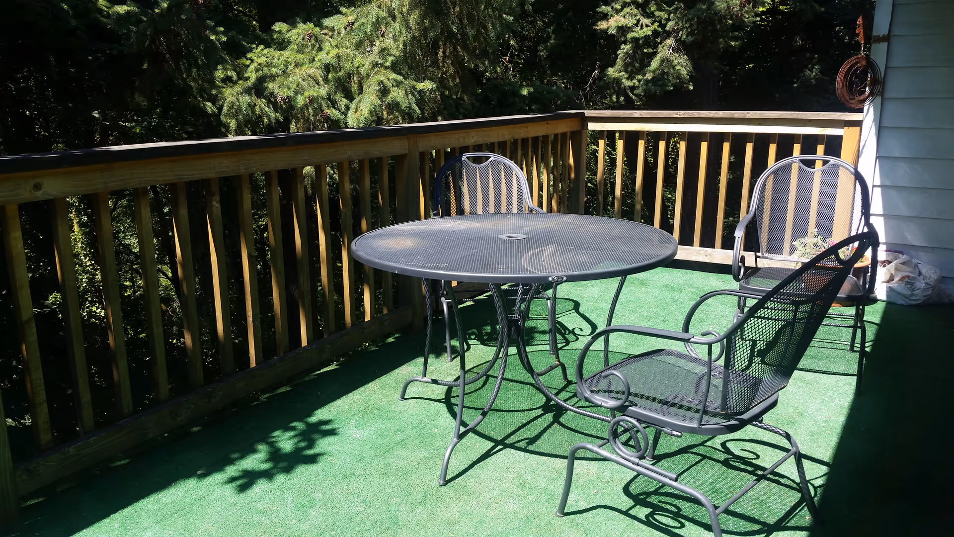 Outdoor patio area with green artificial grass flooring, a round metal table, and three metal chairs. The patio is surrounded by a wooden railing and shaded by trees in the background.
