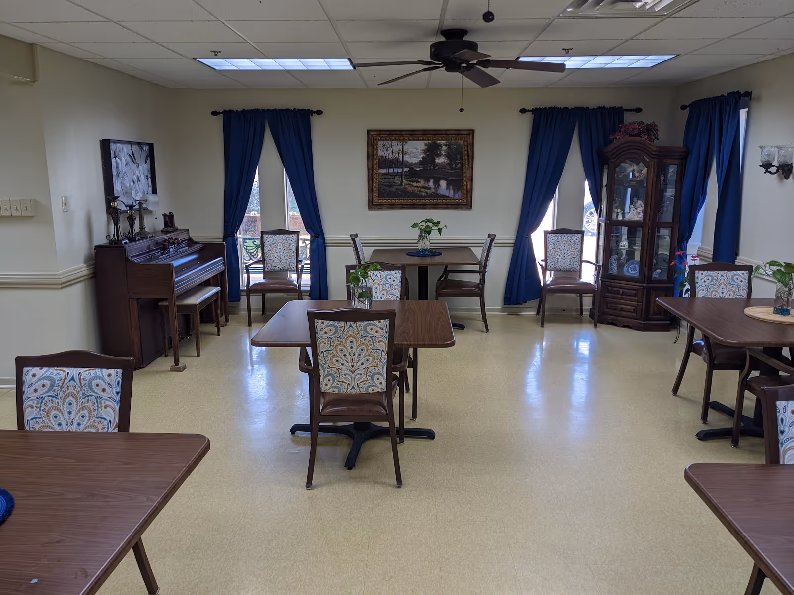 A dining room with several wooden tables and chairs featuring patterned upholstery. The room has cream-colored walls and a light yellow floor. There are three windows with blue curtains, a ceiling fan, a piano with decorative items on top, a framed landscape painting on the wall, and a glass-fronted cabinet displaying dishes and decorative items.
