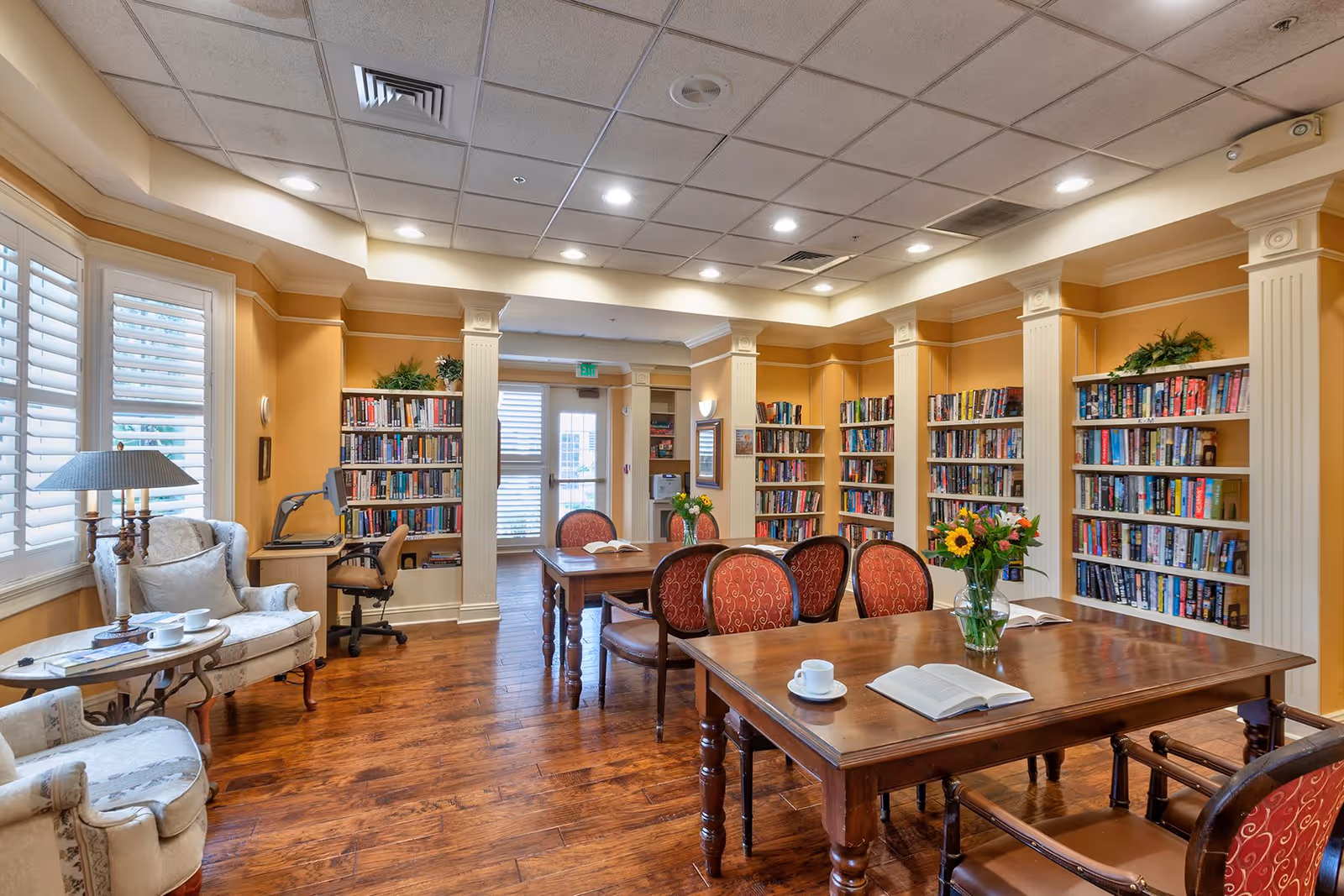 A cozy library room with wooden floors, multiple bookshelves filled with books, and two wooden tables with red cushioned chairs. There are flowers in vases on the tables, an open book, and a cup and saucer. To the left, there are two upholstered armchairs with a small round table and a lamp. A desk with a computer chair is positioned near the window with white shutters. The room has warm yellow walls and a ceiling with recessed lighting.